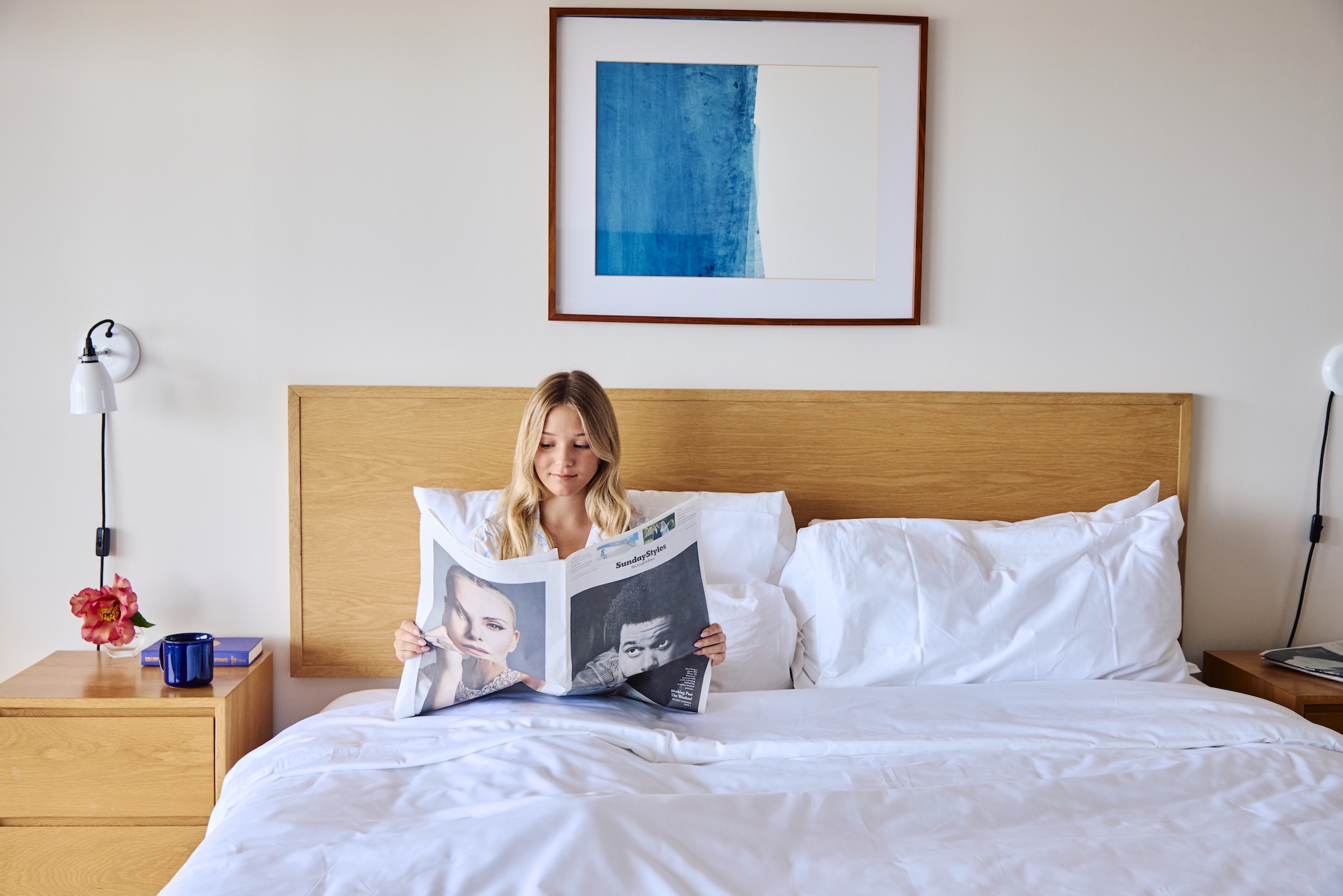 A woman reading a newspaper while in bed with coffee.