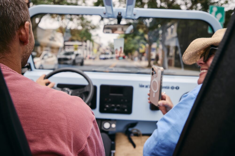A woman talking a photo of a guy driving a Moke in Greenport, NY.