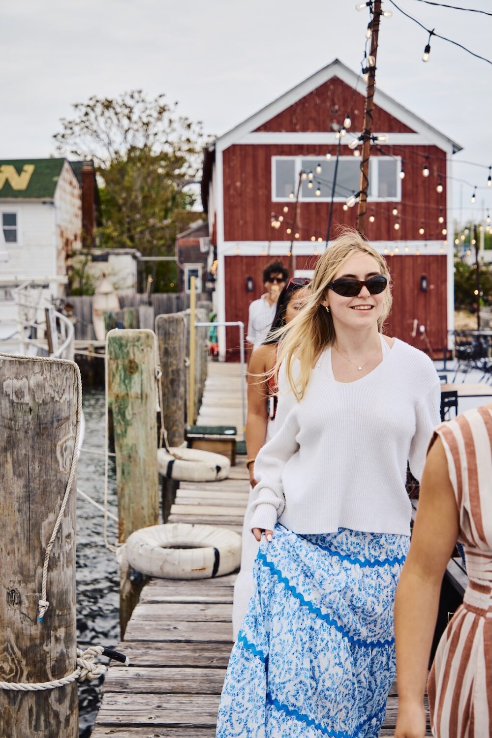 A woman walking down the pier, wearing a white sweater and blue skirt.